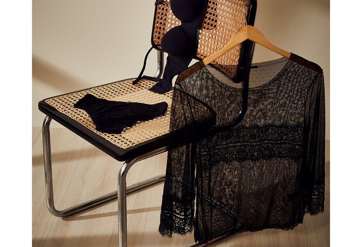 Blusa de renda preta pendurada em um cabide e calcinha e sutiã sob cadeira de madeira. Imagem para ilustrar conteúdo sobre g-string.